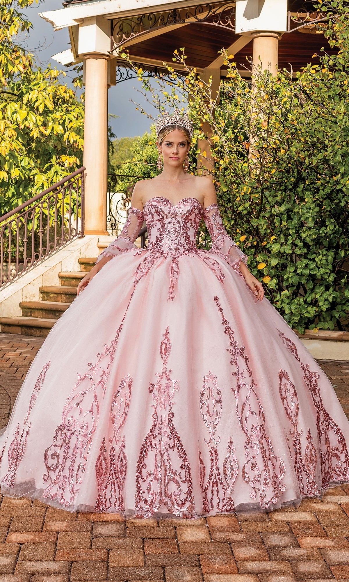 A woman in the Quinceanera Dress 1861 By Dancing Queen, a blush pink, beaded strapless sweetheart gown, with a tiara, stands outdoors on a brick path surrounded by lush foliage and a pavilion.